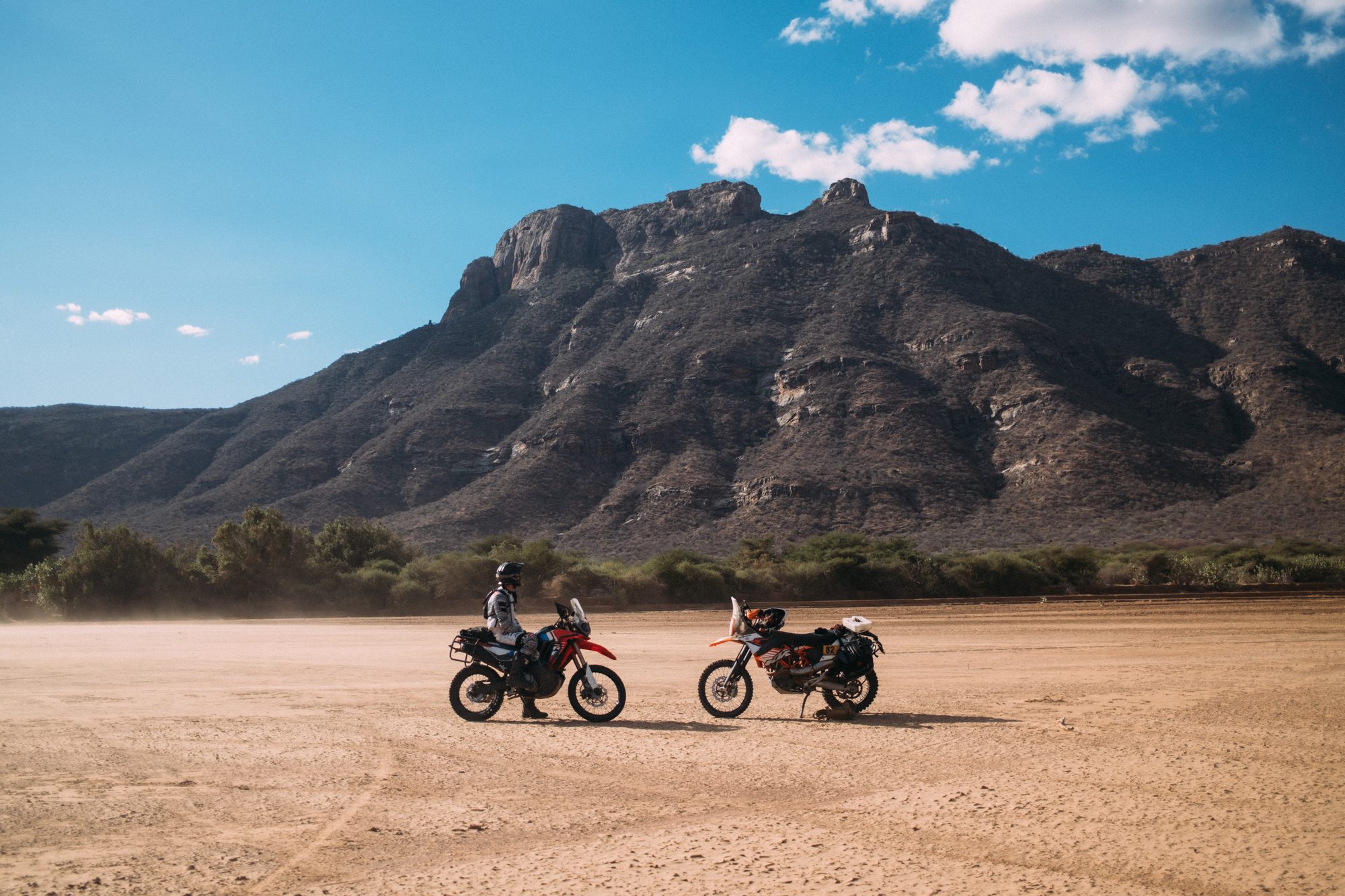 Two motorbikes on a dry riverbed below a long rock wall, riders beside them