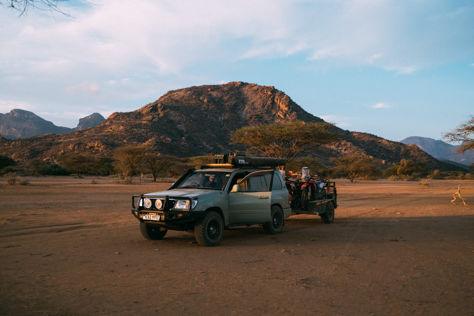 A support vehicle on a dry riverbed below a red rock mountain