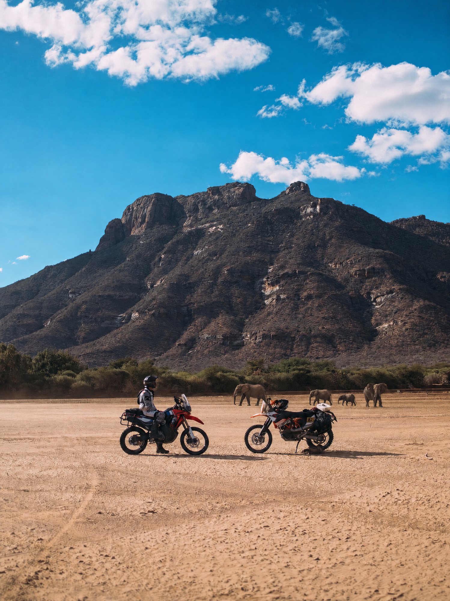 Two motorbikes on a dry riverbed beneath a rocky mountain, elephants walking in the distance